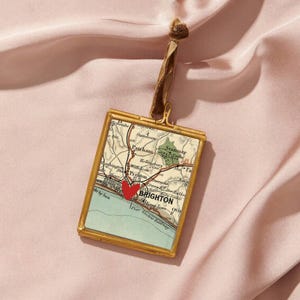 Può includere: Un ciondolo rettangolare dorato con una mappa di Brighton, in Inghilterra, e un cuore rosso che indica una località. Il ciondolo è appeso a un nastro marrone su uno sfondo rosa.