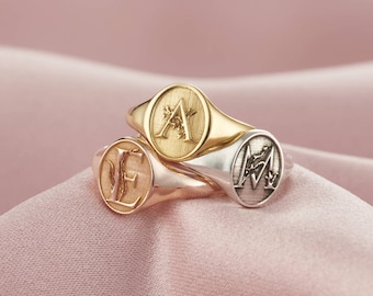 May include: Three gold and silver signet rings with engraved letters A, E, and M. The rings are stacked on top of each other on a pink fabric background.