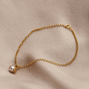 May include: A delicate gold chain bracelet with a single pearl charm. The chain is a simple, linked design and the pearl is white and round.