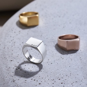 Könnte beinhalten: Drei quadratische Siegelringe, einer in Silber, einer in Gold und einer in Roségold. Der silberne Ring trägt die Inschrift "S.A.F." auf der Oberseite.