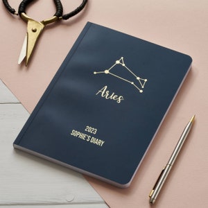 May include: A navy blue diary with a gold foil design of the Aries constellation and the text "Aries" and "2023 Sophie's Diary".