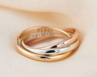 May include: Three interlocking gold rings with a rose gold band, a silver band, and a yellow gold band. The rings are engraved with the name "HENRY" and the date "08.10.22". Two small diamonds are set in the rings.