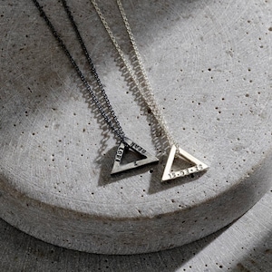 May include: Two silver triangle pendant necklaces with chain links. One necklace has a dark finish, the other is silver. The pendants have engraved text, including names and dates. The necklaces are displayed on a gray, textured surface.
