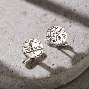 May include: A pair of round silver-toned cufflinks featuring engraved black line maps of city street grids. One cufflink is labeled with the text Brighton, and the other cufflink is labeled with the text Nottingham, both in a clean sans-serif font.