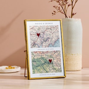 May include: A gold framed map print with two sections. Each section shows a vintage map with a red heart marking a location. The top section reads "Maisie & George" and "Engaged 20.07.2023". The bottom section reads "Married 30.05.2024".