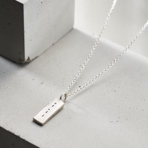 Puede incluir: Un collar de plata con un colgante rectangular que presenta una serie de puntos. El collar está sobre una superficie gris.