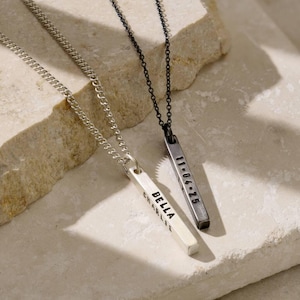 Pode incluir: Dois colares de barra personalizados. Um é prateado com o nome "BELLA CHARLIE" gravado. O outro é cinza escuro com a data "11-04-25" gravada. Ambos os colares têm uma corrente.