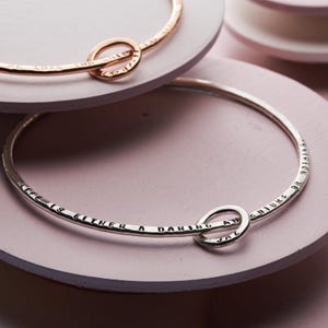 Könnte beinhalten: Silbernes Armreif-Armband mit einem Knotenring-Anhänger. Das Armband ist mit dem Text "Das Leben ist entweder ein wagemutiges Abenteuer oder gar nichts" graviert.