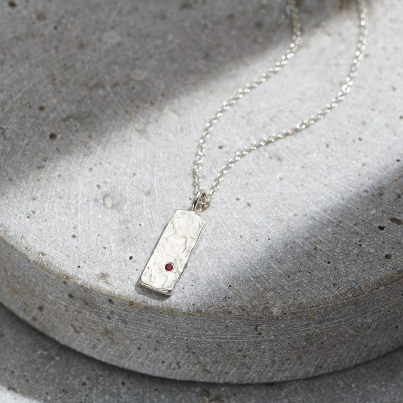 May include: A silver necklace with a textured rectangular pendant featuring a small red gemstone.