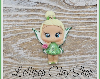 May include: A handcrafted fairy figurine with blonde hair, blue eyes, and a green dress with glitter accents. The fairy holds a pink rose and has white and green glitter wings. The text "Lollipop Clay Shop" is at the bottom.
