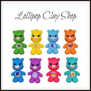 May include: A collection of eight colorful clay bear figurines. Each bear is a different color, including green, yellow, blue, orange, purple, and pink. The bears have heart-shaped noses and symbols on their bellies. The text "Lollipop Clay Shop" is at the top.