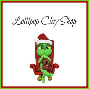 May include: A green character wearing a Santa hat, seated in a red chair, holding a brown dog. The character has a grumpy expression. The text "Lollipop Clay Shop" is above the character, all within a red and green border.