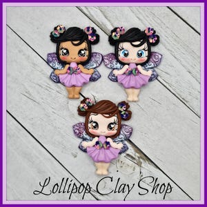 May include: Three handmade fairy figurines with black, brown, and tan hair, wearing lavender dresses and iridescent wings. Each fairy has a flower in their hair and is smiling. The text "Lollipop Clay Shop" is at the bottom.