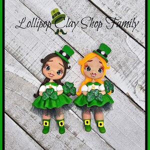 May include: Two clay figurines of leprechaun girls. One has brown hair, the other has blonde hair. Both wear green dresses, hats, and boots, and hold shamrocks. The text "Lollipop Clay Shop Family" is above the figurines.