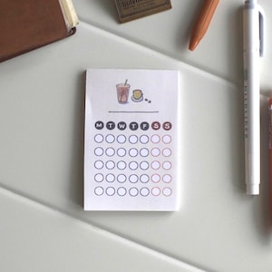 May include: A small, white notepad with a calendar layout, featuring a coffee illustration at the top. The days of the week are abbreviated, and there are circles for marking dates. A brown notebook, pens, and other stationery items are nearby.
