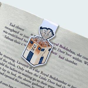 May include: A bookmark featuring a cartoon illustration of a loaf of bread in a clear plastic bag. The bookmark has a white tab and is placed on a book with text.