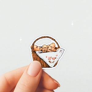 May include: A sticker depicting a wicker basket holding several loaves of bread. The basket has a handle and is lined with a white cloth decorated with pink floral patterns. The bread is golden brown with detailed markings.