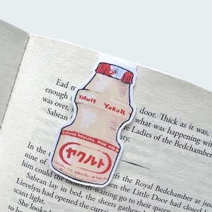 May include: A bookmark shaped like a Yakult bottle, featuring the brand name in English and Japanese. The bookmark is placed in an open book, with text visible on the pages. The bottle is beige with a red cap.