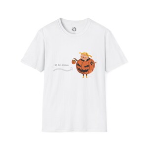 Puede incluir: Camiseta blanca con una ilustración de dibujos animados de una persona con un disfraz de calabaza sosteniendo una bolsa de truco o trato. El texto "tis the season" está impreso en la camiseta. El diseño es naranja, marrón y amarillo.