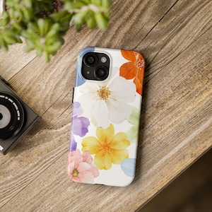 Puede incluir: Una funda blanca para smartphone con un diseño floral colorido. La funda muestra flores en tonos naranja, amarillo, rosa, morado y blanco. La funda está sobre una superficie de madera.