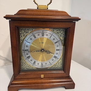 May include: A rectangular wooden clock with a gold-colored face and Roman numerals. The clock has a brass handle on top and a decorative gold border around the face. The clock face reads "HOWARD MILLER".