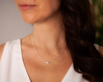 May include: A silver chain necklace with a small, square pendant. The necklace is worn by a person with long, dark hair.