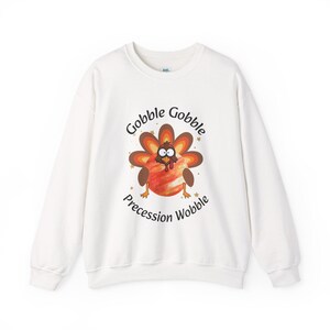 May include: White sweatshirt with a colorful turkey graphic. The turkey has a brown and orange feathered body, a red and orange belly, and the text "Gobble Gobble Precession Wobble" in a playful font. A fun, festive design.