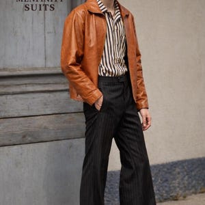 May include: A man wearing a brown leather jacket over a striped shirt and black pinstripe pants. The jacket is a warm brown color. The shirt has vertical black and white stripes. The pants are flared. The text "MENFINITY SUITS" is visible.