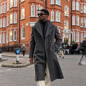 May include: A charcoal gray double-breasted overcoat with wide lapels and buttons, worn over a black turtleneck and white trousers. The garment features a textured fabric and is paired with a dark patterned scarf and black sunglasses.