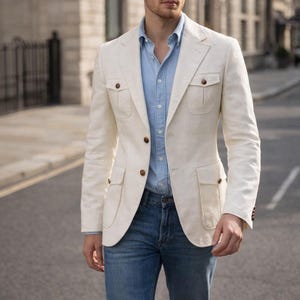 Peut inclure: Veste en lin blanc cassé avec quatre poches avant et des boutons marron, portée sur une chemise bleu clair et un jean bleu. La veste a une coupe classique et ajustée. Le texte "MENFINITY SUITS" est visible.