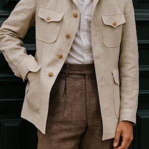 Puede incluir: Chaqueta de lino beige con cuatro bolsillos delanteros y botones de madera, combinada con pantalones marrones. La chaqueta se lleva sobre una camisa blanca. El logotipo "MENFINITY SUITS" es visible.