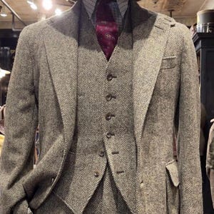 May include: A three-piece suit in a brown and grey herringbone tweed fabric, featuring a notched lapel jacket, a five-button vest, and matching trousers. The outfit includes a light blue and white checkered shirt, a dark red paisley tie, and a brown hat.