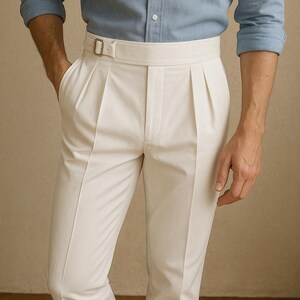 May include: Cream-colored pleated trousers with a high waist and a matching belt. The trousers are paired with a light blue button-down shirt. The image shows a close-up of the pants and shirt, showcasing the details of the fabric and design.