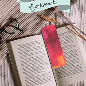 Op de afbeelding: Een rechthoekige boekenlegger met een levendig kleurverloop van roze, rood en oranje, rustend op een open boek. De boekenlegger heeft een kleine, gestileerde handtekening in de onderste hoek. Daarnaast liggen een bril met donker montuur en een tarwestengel.