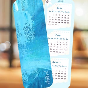 Op de afbeelding: Een verticale bladwijzerkalender voor 2026 met een gestructureerd blauw abstract geschilderd ontwerp op de voorzijde en een lichtblauwe achtergrond met witte papierachtige blokken voor de maanden juni, juli en augustus.