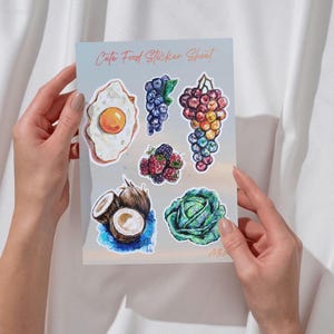 Op de afbeelding: Een stickervel met de titel Cute Food Sticker Sheet met aquarel-stijl illustraties van een gebakken ei, bosbessen, een tros veelkleurige druiven, frambozen, bramen, gehalveerde kokosnoten en een krop groene kool.