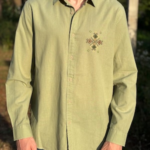 Puede incluir: Una camisa de manga larga de color verde oliva claro con cuello en punta y una hilera vertical central de botones a juego. Un peque&ntilde;o y complejo patr&oacute;n de bordado geom&eacute;trico en tonos verdes, marrones y amarillos se sit&uacute;a en el pecho izquierdo.