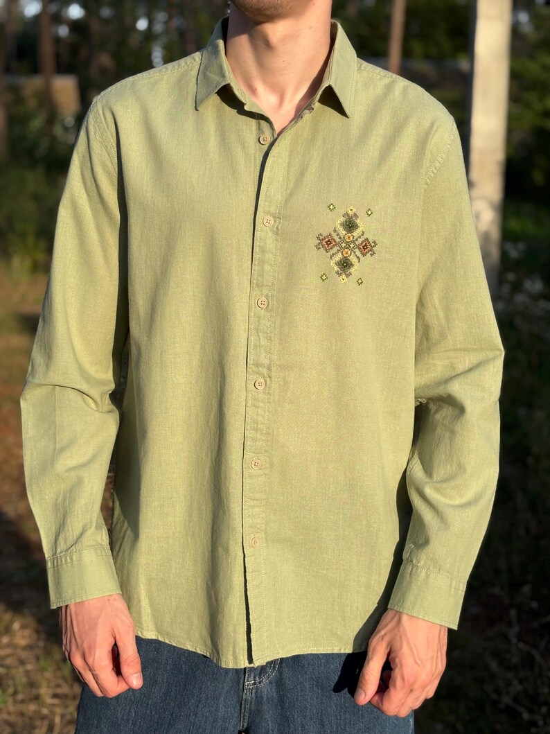 Puede incluir: Una camisa de manga larga de color verde oliva claro con cuello en punta y una hilera vertical central de botones a juego. Un peque&ntilde;o y complejo patr&oacute;n de bordado geom&eacute;trico en tonos verdes, marrones y amarillos se sit&uacute;a en el pecho izquierdo.