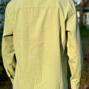 Puede incluir: Una camisa de manga larga con botones en un color verde lima claro. La camisa presenta un cuello est&aacute;ndar, un canes&uacute; en la espalda y pu&ntilde;os abotonados en las mangas. La tela tiene una textura visible y la camisa se lleva sobre unos vaqueros azules.