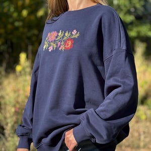 May include: A navy blue crewneck sweatshirt featuring an embroidered floral design on the chest. The embroidery consists of red, pink, and yellow flowers with green leaves. The sweatshirt has long sleeves with ribbed cuffs and a ribbed neckline.
