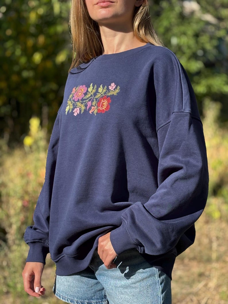 May include: A navy blue crewneck sweatshirt featuring an embroidered floral design on the chest. The embroidery consists of red, pink, and yellow flowers with green leaves. The sweatshirt has long sleeves with ribbed cuffs and a ribbed neckline.