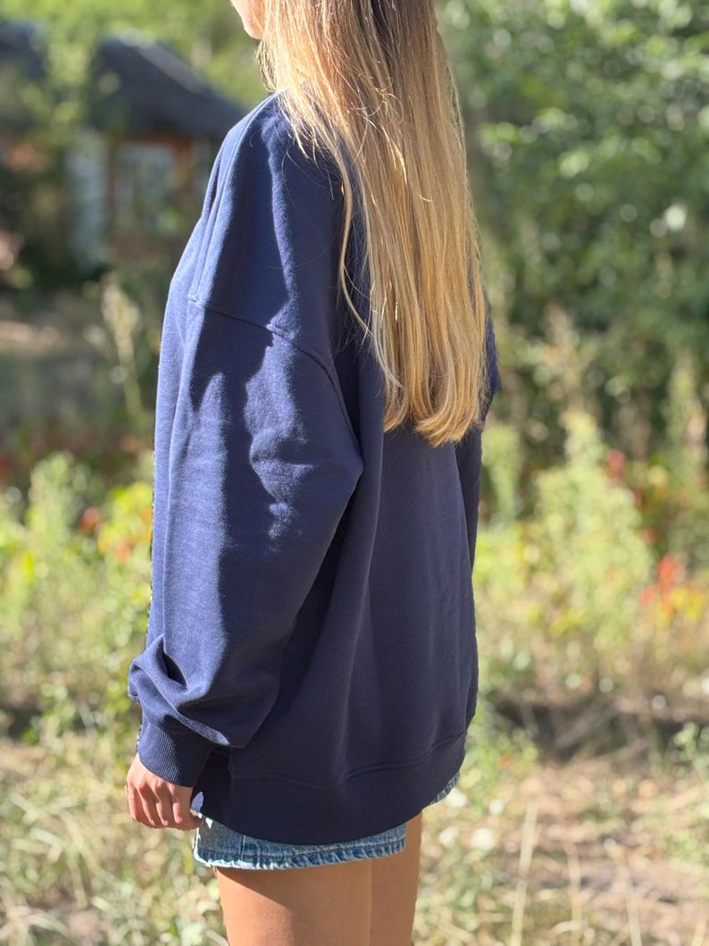 May include: A navy blue long-sleeved sweatshirt with a relaxed fit, featuring ribbed cuffs and a ribbed hemline. The garment is worn over light blue denim shorts, showcasing a simple and functional design with a solid dark blue colour throughout.