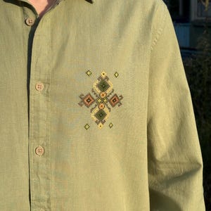 Puede incluir: Una camisa de manga larga abotonada en color verde salvia con un peque&ntilde;o e intrincado bordado de punto de cruz en el pecho izquierdo. El bordado consiste en formas geom&eacute;tricas de diamante en tonos de hilo verde oscuro, amarillo y marr&oacute;n.