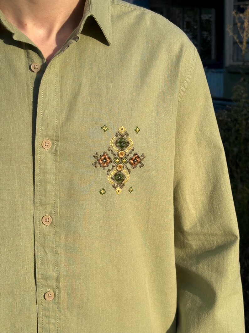 Puede incluir: Una camisa de manga larga abotonada en color verde salvia con un peque&ntilde;o e intrincado bordado de punto de cruz en el pecho izquierdo. El bordado consiste en formas geom&eacute;tricas de diamante en tonos de hilo verde oscuro, amarillo y marr&oacute;n.