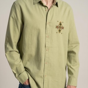 May include: A long-sleeved button-down shirt in a light olive green color. The shirt features a classic collar and a small, intricate geometric embroidery pattern in shades of yellow, brown, and green located on the upper left chest area.