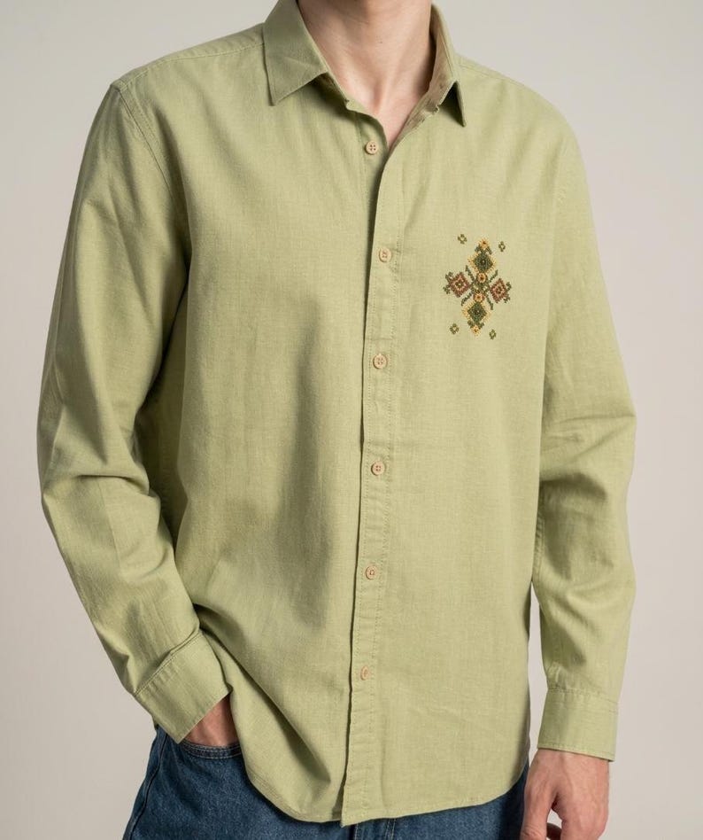 Puede incluir: Una camisa de manga larga con botones en color verde oliva claro. La camisa presenta un cuello cl&aacute;sico y un peque&ntilde;o y complejo patr&oacute;n geom&eacute;trico bordado en tonos amarillos, marrones y verdes situado en la parte superior izquierda del pecho.