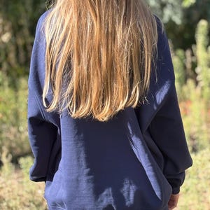 May include: A navy blue long-sleeved sweatshirt with a crew neckline and ribbed cuffs and hem, paired with light blue denim shorts. The sweatshirt features a loose, relaxed fit and is worn over the shorts, covering the upper portion of the legs.