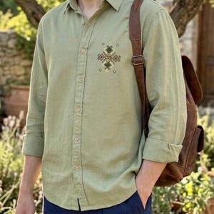 Puede incluir: Una camisa de manga larga color verde salvia con un peque&ntilde;o patr&oacute;n geom&eacute;trico bordado en tonos marr&oacute;n, verde y crema en el pecho izquierdo, acompa&ntilde;ada de una mochila de cuero marr&oacute;n con una sola correa visible.
