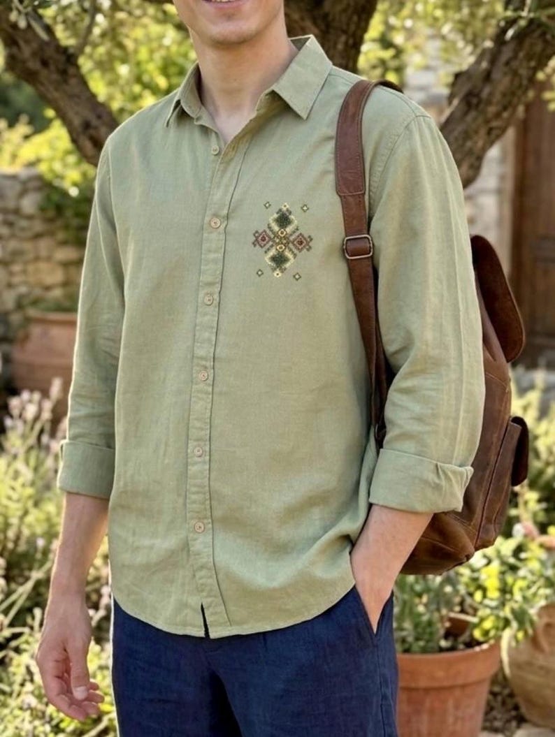 Puede incluir: Una camisa de manga larga color verde salvia con un peque&ntilde;o patr&oacute;n geom&eacute;trico bordado en tonos marr&oacute;n, verde y crema en el pecho izquierdo, acompa&ntilde;ada de una mochila de cuero marr&oacute;n con una sola correa visible.