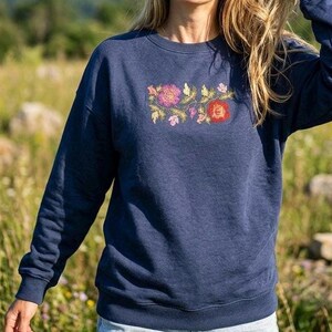 May include: A navy blue long-sleeve sweatshirt featuring a central embroidered design of two large flowers in shades of pink and red, surrounded by smaller green leaves and delicate floral accents, with a ribbed crew neckline and cuffs.